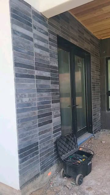 Charcoal linear stacked stone cladding around a black-framed glass entry door with a wood soffit above