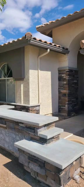 Stone-faced outdoor kitchen island with concrete countertops beside a stone column on a tile-roofed patio